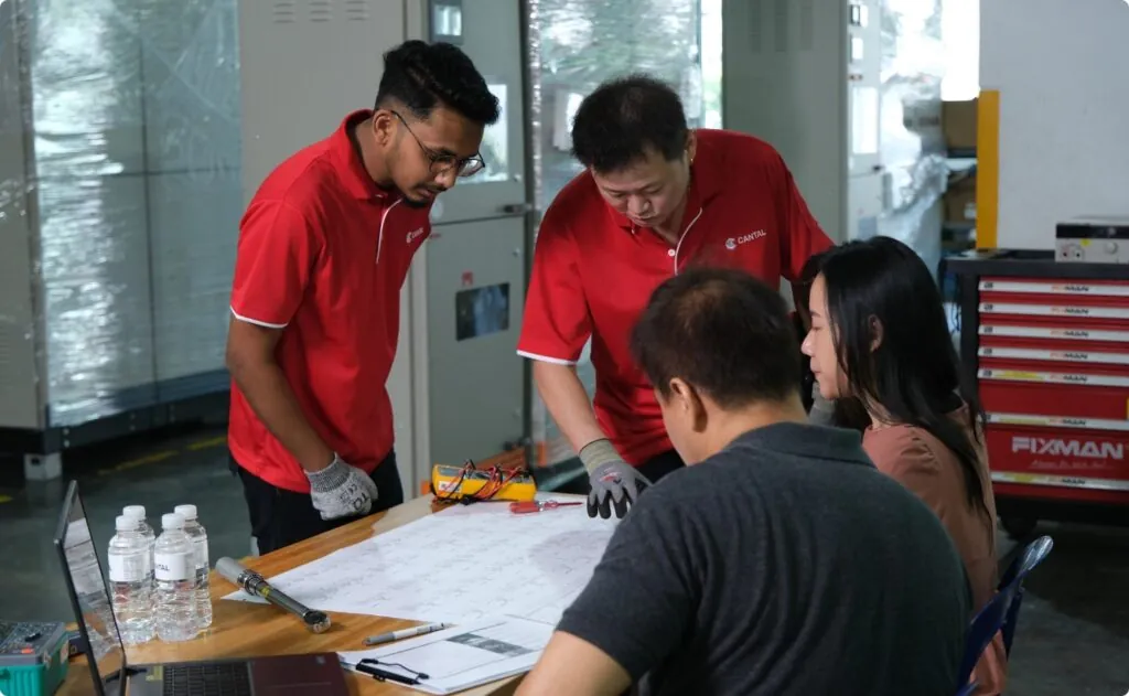 A team of four professionals, including engineers and technicians in red Cantal shirts, intently reviewing electrical blueprints and schematics on a table in an industrial workshop setting. Various tools, a laptop, and water bottles are also on the table.