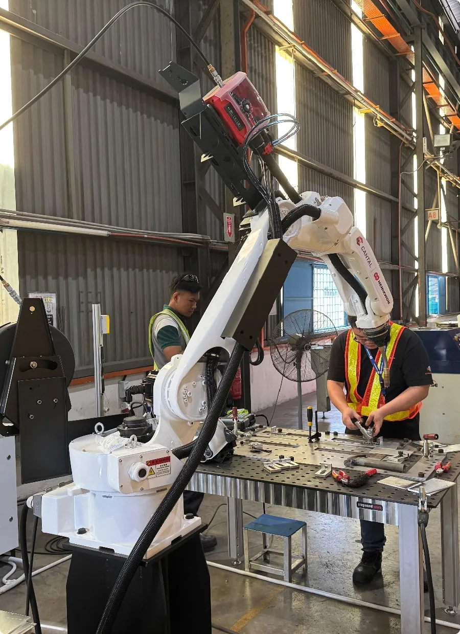 White industrial robotic arm and two workers fabricating metal components at a workbench in a factory setting.