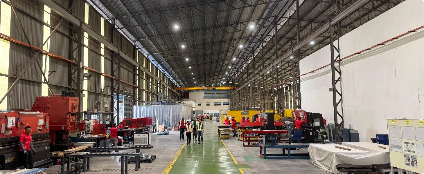 A bustling industrial factory floor with red machinery, workers in red shirts, and a group in safety vests walking down a central green aisle.
